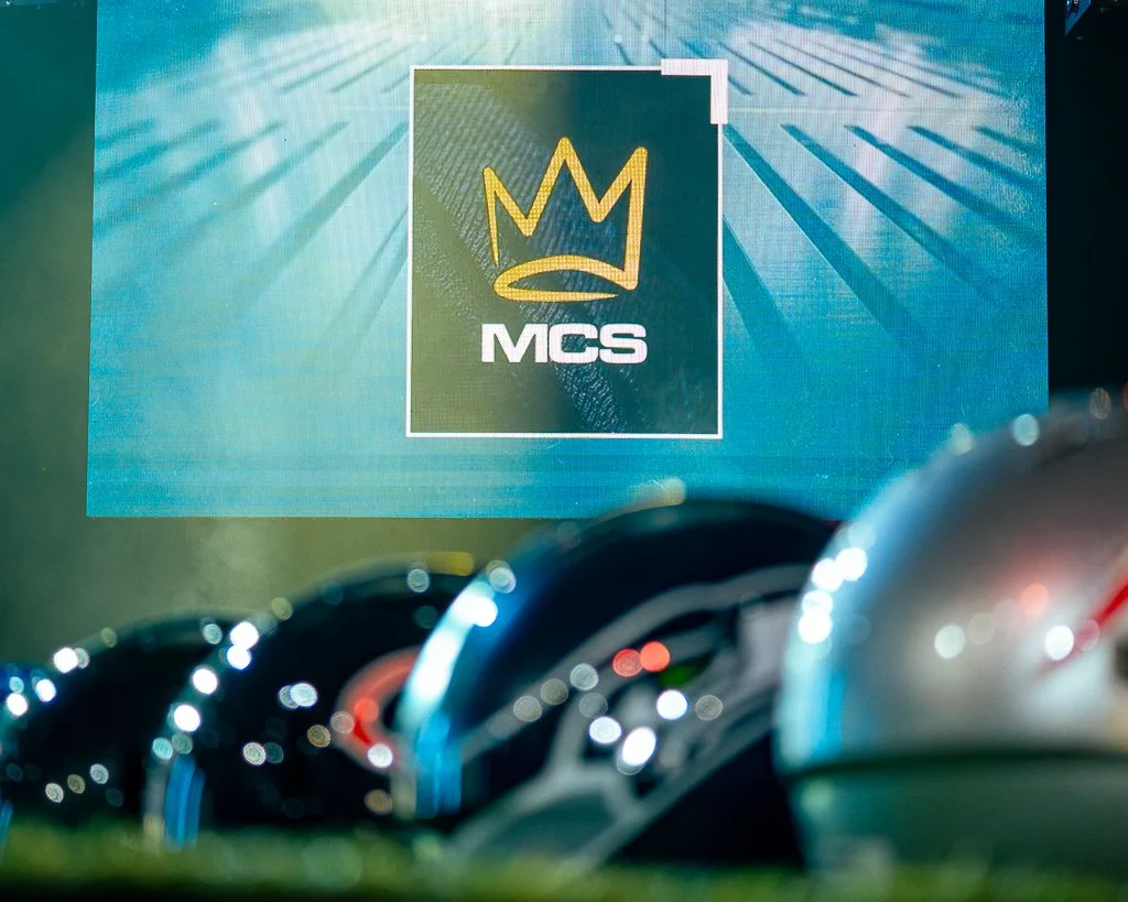 A digital screen displaying the logo of MCS, featuring a gold crown symbol above the white letters 'MCS', with a background of blue and teal rays.
