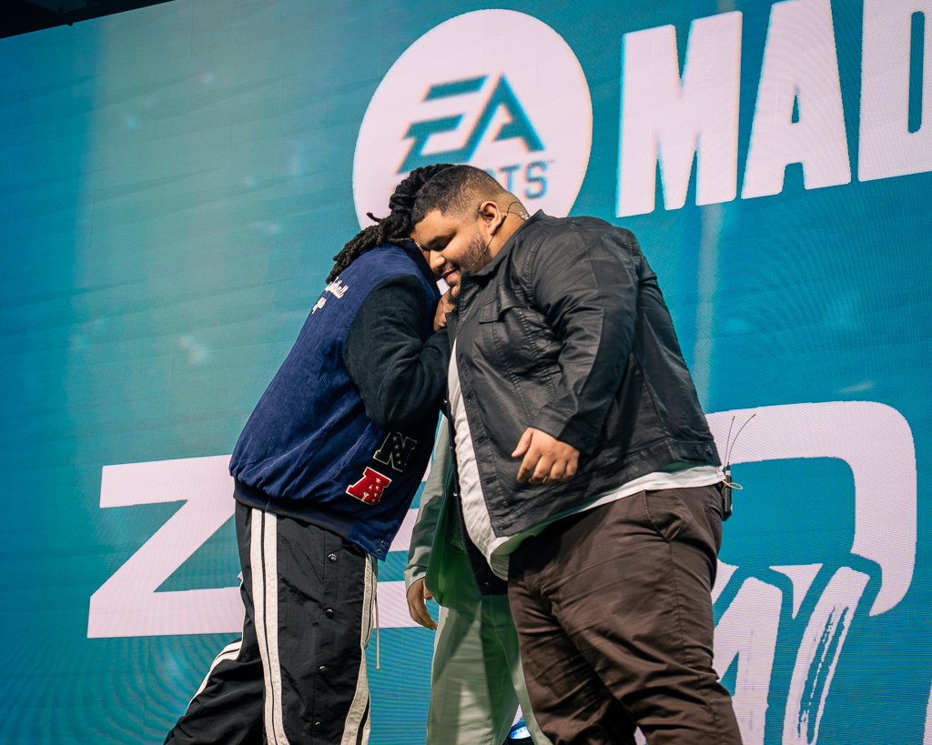 Two men sharing a moment on stage in front of a large EA Sports Madden NFL logo backdrop, hugging each other with closed eyes.
