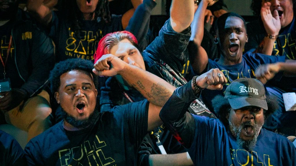A group of diverse young adults showing excitement and passion, with some raising fists and shouting, at an event or gathering.