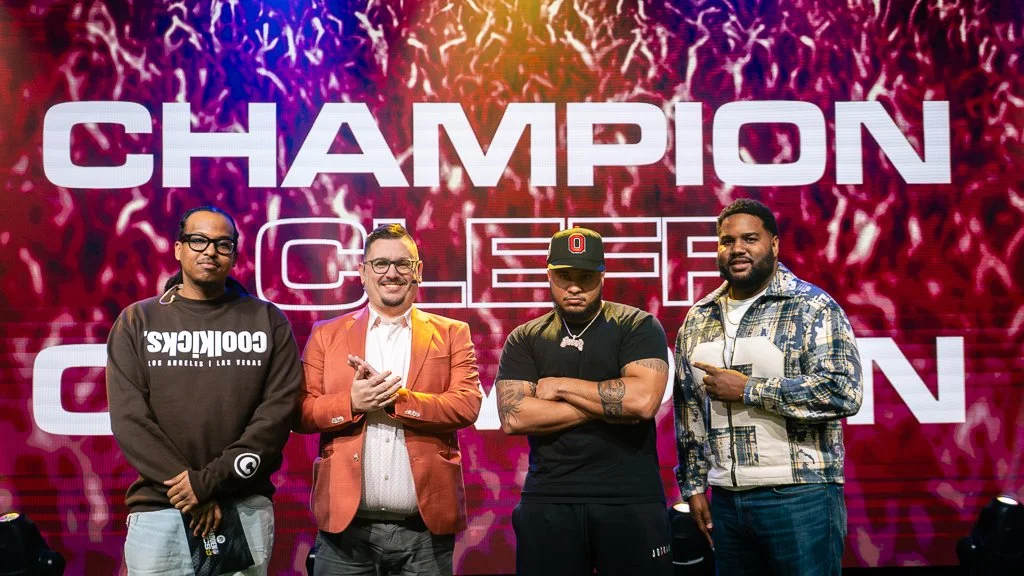 Four men standing in front of a large screen that reads 'Champion Crew'. The men are dressed casually, and the background has a vibrant, fiery design.