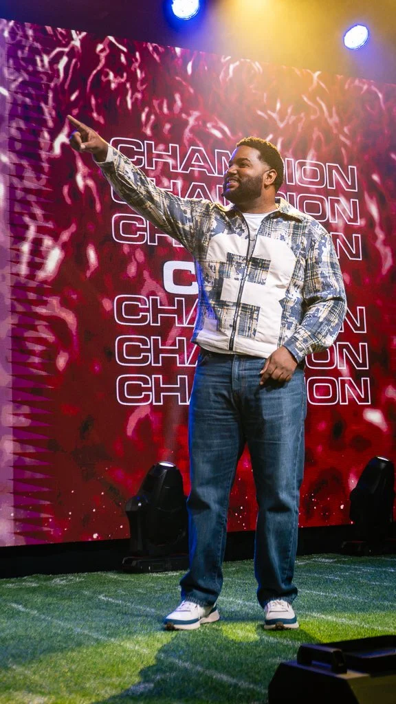 A man standing on stage pointing upward with his right hand, smiling, with a large LED screen behind him displaying the word "CHAMPION" repeatedly in red and white text. The stage has green artificial grass and stage lights are overhead.