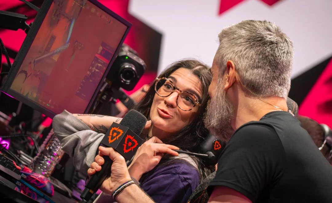 Content creator being interviewed at Valorant North America playoffs holding a microphone at a gaming event, with a computer monitor and gaming equipment visible.