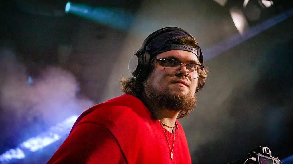 A young man with long hair, glasses, and a beard, wearing a black backward cap, headphones, and a red shirt, standing in a dimly lit music or event venue with fog and colorful lights.