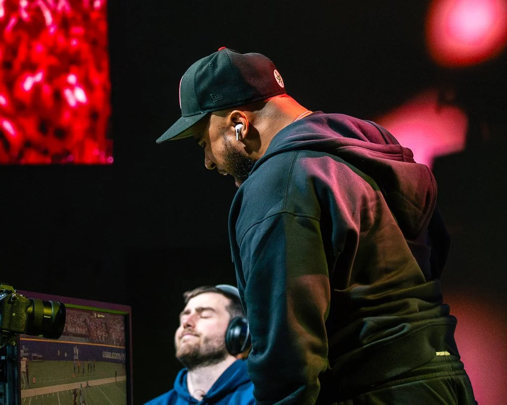 A man wearing a black hoodie and black baseball cap standing near another man with headphones watching a screen showing a sports game.