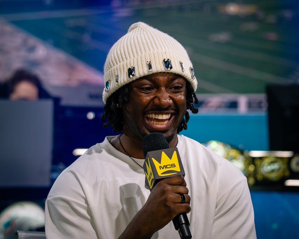 Man with dreadlocks wearing a white beanie with gemstone embellishments, smiling, holding a microphone with a yellow and black MCS logo at an event.