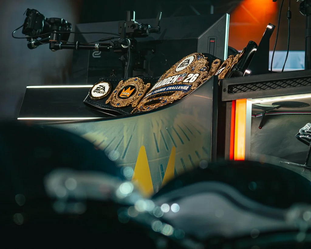 A racing setup with a computer monitor, a steering wheel, and a video game controller on a desk. Several championship belts are draped over the desk, including those with the EA Sports logo and other racing-themed designs. The scene appears to be in 