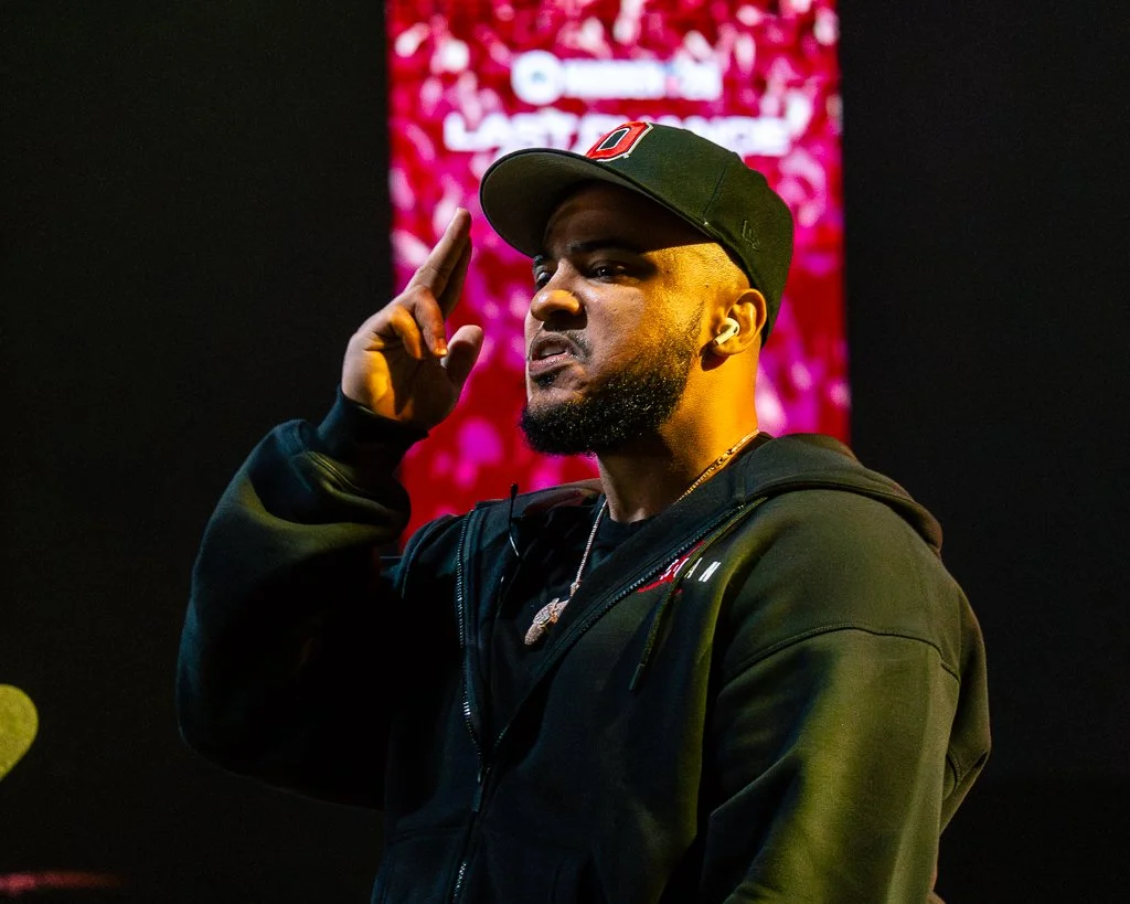A man wearing a black hoodie and a black cap is saluting with his right hand, against a pink and red background with blurred text.