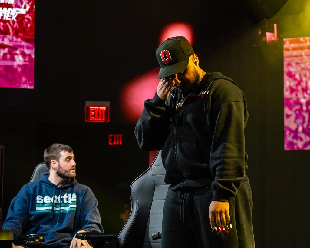 Two men at a gaming event, one sitting at a desk with a computer and the other standing, wearing casual clothing, dark hoodie and cap, with a gaming chair and colorful screens in the background.