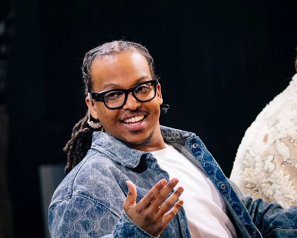 A smiling man with glasses and dreadlocks wearing a denim jacket and white t-shirt, speaking or gesturing, with a dark background.