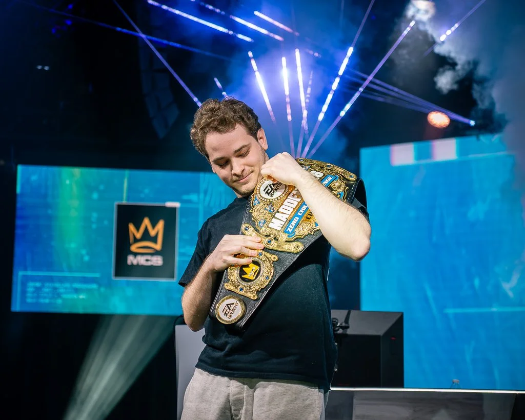 Young man in a black t-shirt holding a championship belt on stage at an esports event with colorful laser lights and a large screen in the background.