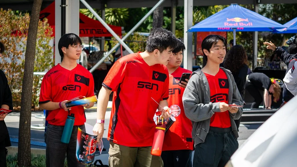 Group of esports fans in sentinels jerseys standing outdoors at the North America Playoffs in LA, with some holding water bottles, pamphlets, and smartphones, at an event with Blue umbrellas and metal scaffolding in the background.
