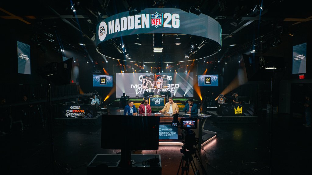 Stage setup for Madden NFL 26 gaming event with a large circular screen displaying the Madden NFL 26 logo, four hosts or commentators seated at a desk, and multiple large screens showing game graphics, in a dark auditorium with focused lighting.