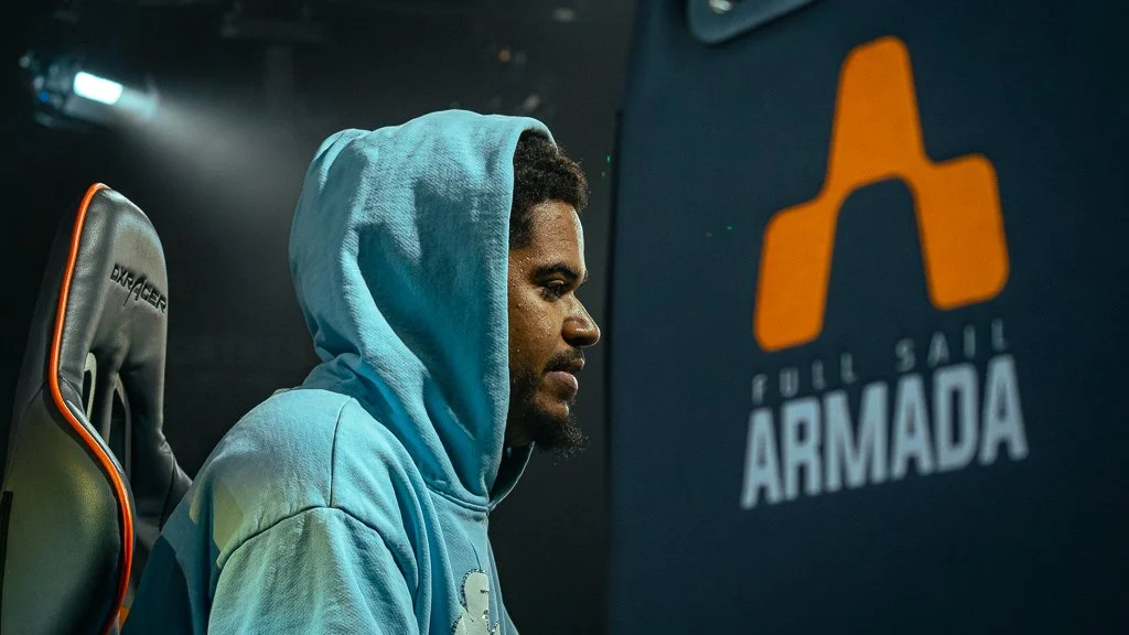 A man in a blue hoodie sitting next to a gaming chair, with a video game logo on a screen that says "Full Sail Armada".