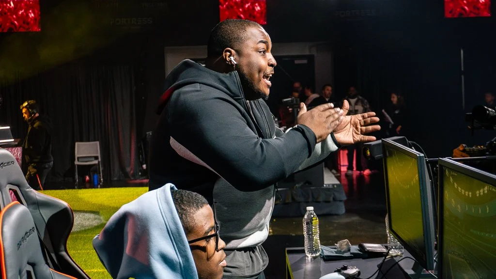 A man clapping and celebrating in front of gaming screens at an esports event, with other people in the background.