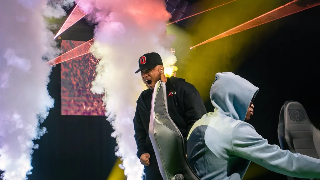 One person wearing a black cap and hoodie is standing and shouting with clenched fist, behind a seated person with an open hoodie and glasses, in a setting with smoke and laser lights.