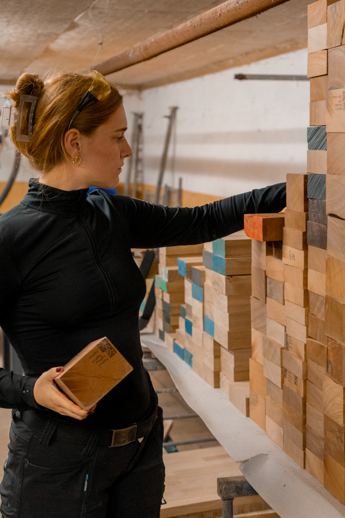 A woman in black clothing and safety glasses inspecting wooden blocks in a workshop.