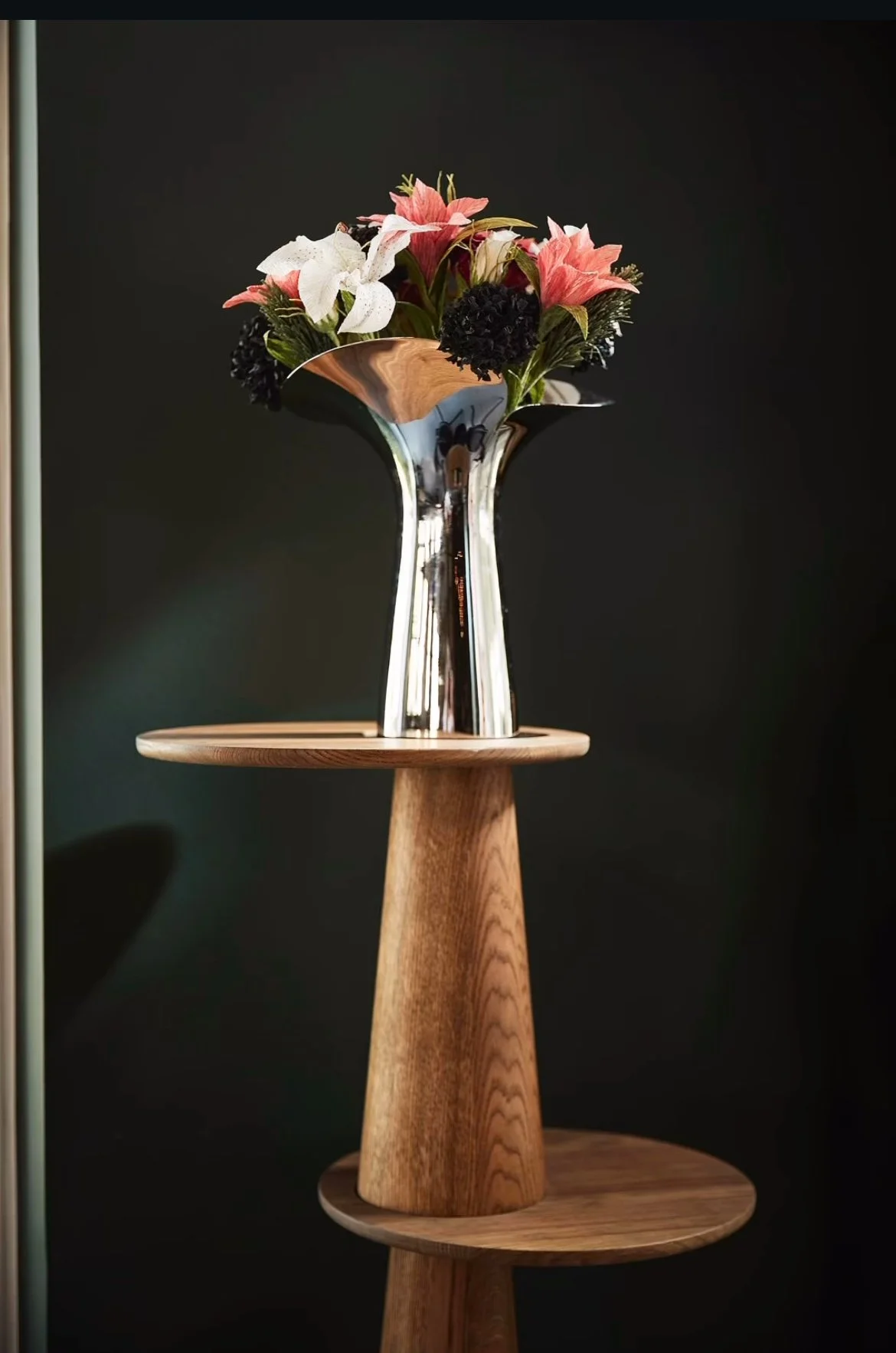 A tall, wooden side table with two circular platforms, on top of which is a shiny metallic vase holding a bouquet of pink and white flowers with some dark accents against a dark background.