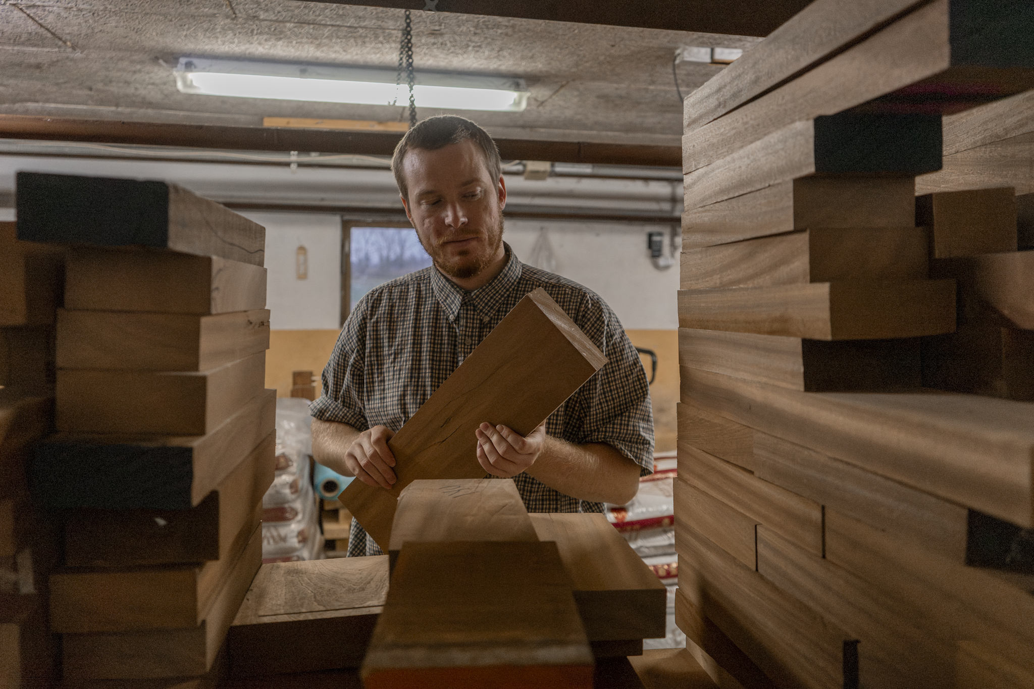 Man inspecting a piece of wood in a woodworking shop, surrounded by stacks of finished lumber.
