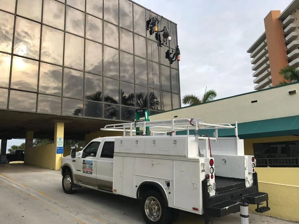 Workers are cleaning the glass windows of a building using harnesses and ropes, while a service truck with equipment is parked nearby in an urban area.