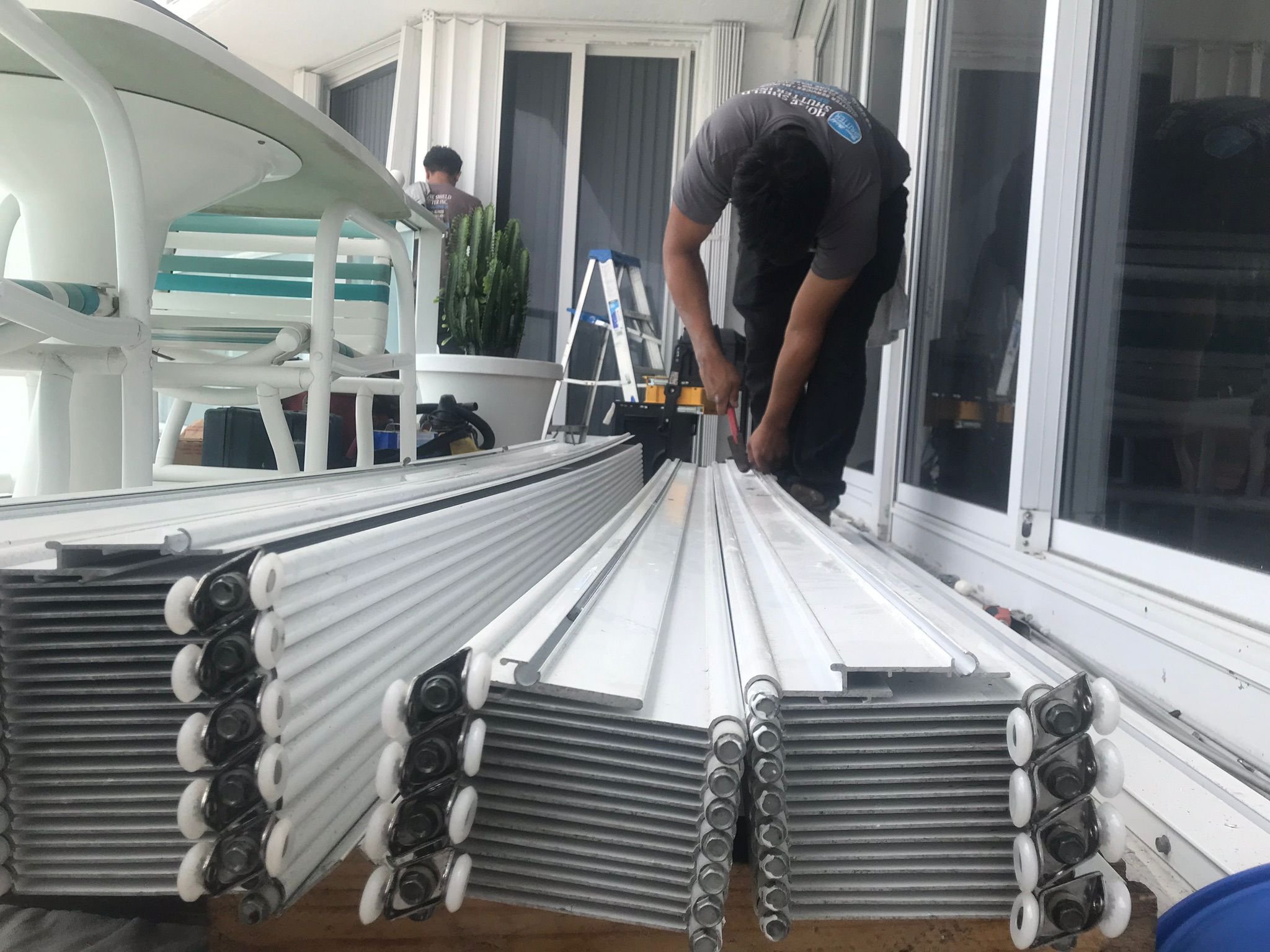 A person working on assembling or repairing white metal or plastic parts on a balcony or patio. There are tools and equipment nearby, including a ladder and a large pot with a cactus plant in the background.