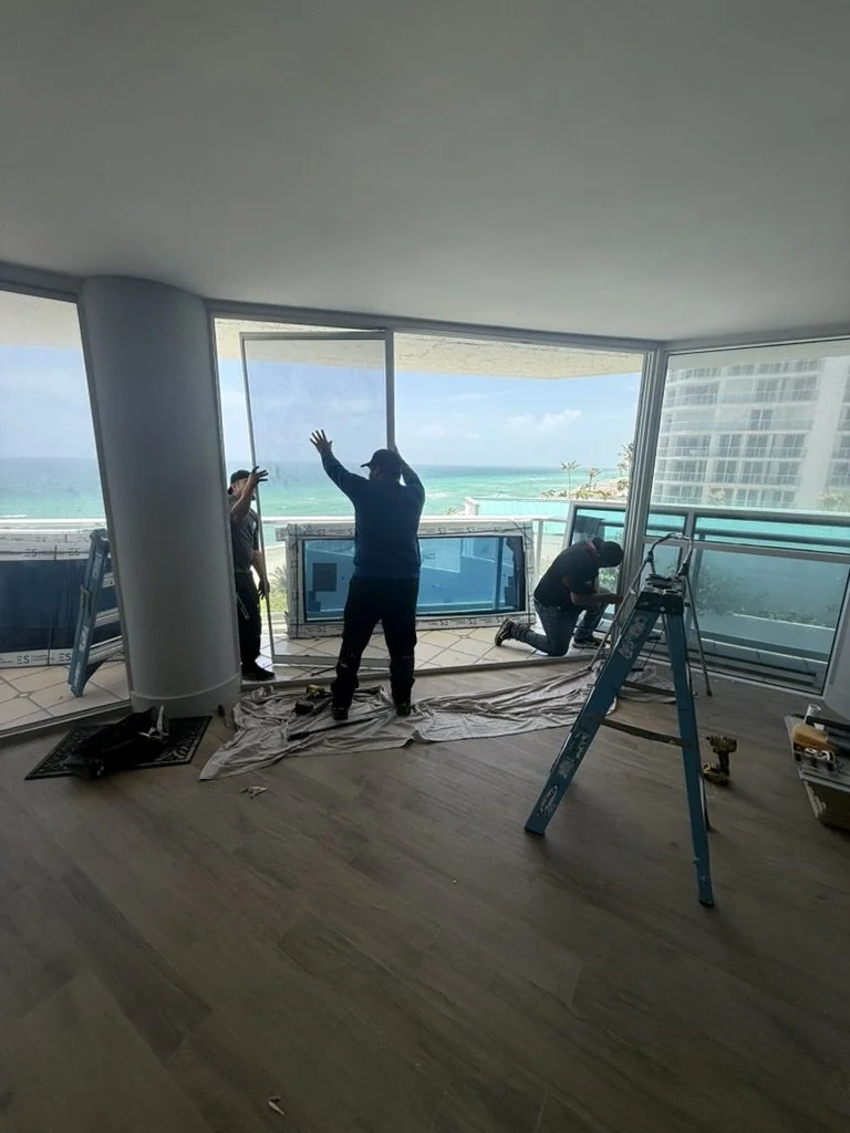 Three workers installing a large flat-screen TV or monitor in a high-rise apartment with floor-to-ceiling windows overlooking the ocean.