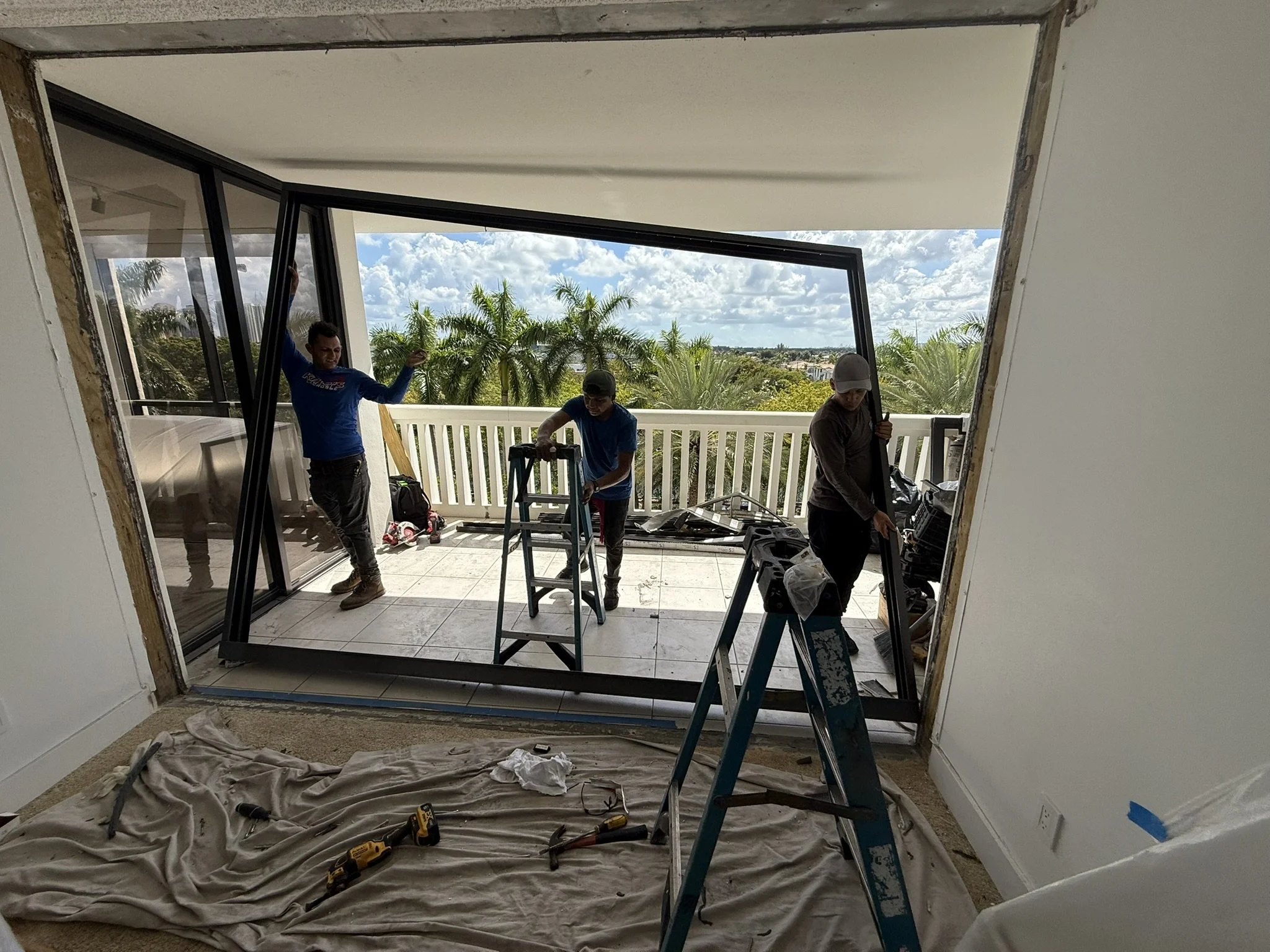 Three workers install a large sliding glass door on a balcony with a view of palm trees and blue sky.