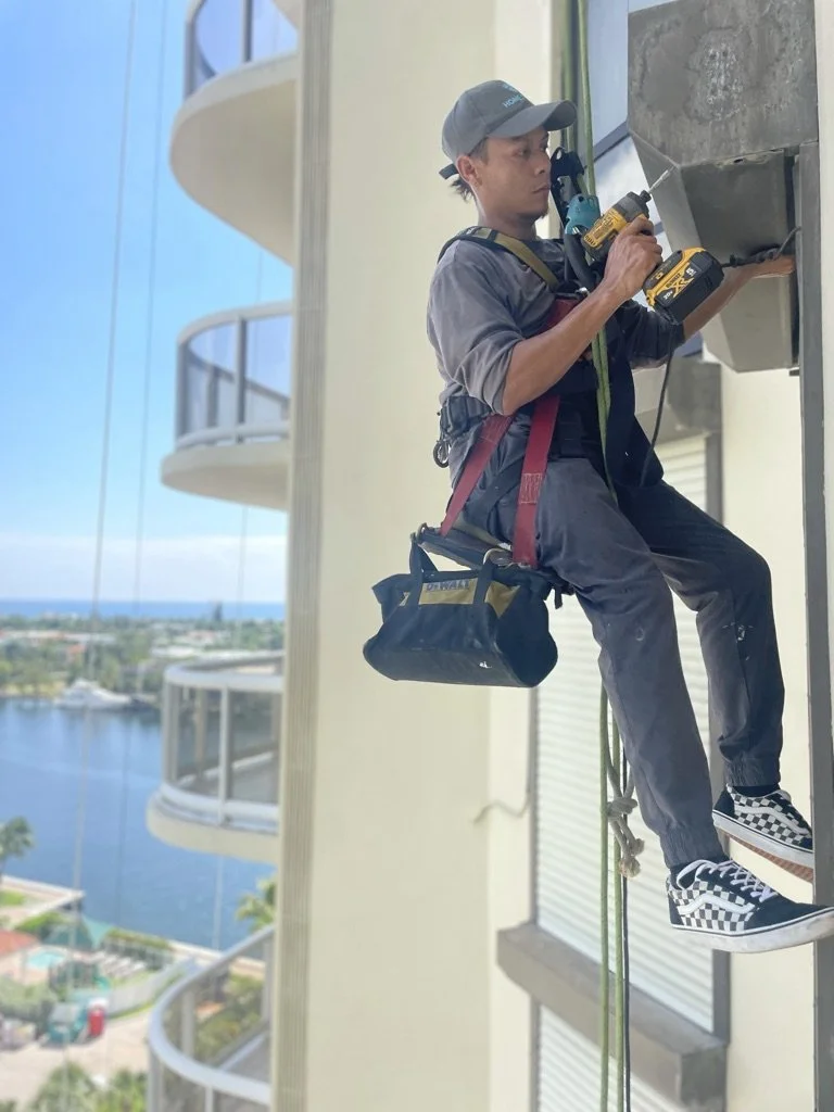 A worker in safety gear, including a harness and a helmet, is cutting or drilling into the exterior wall of a high-rise building with a cordless drill.