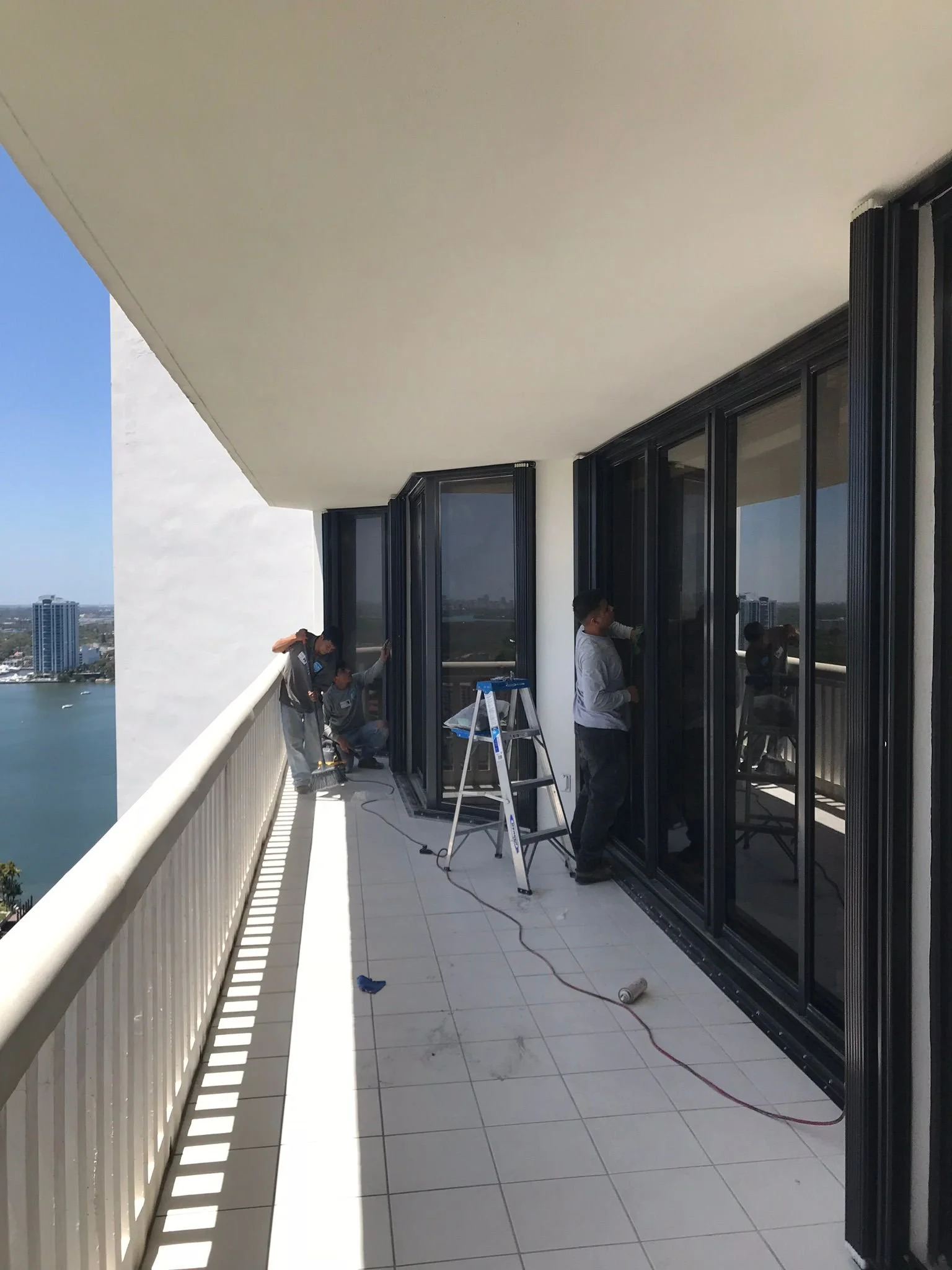 Construction workers installing black-framed glass doors on a balcony with city and water view in the background.