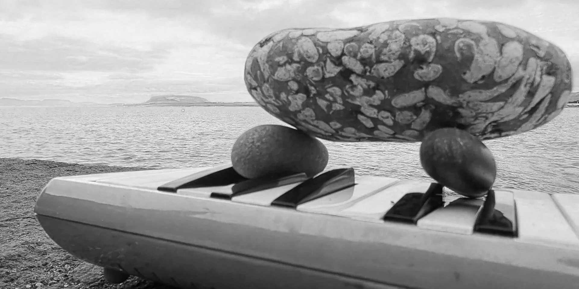 Stones resting on melodica keys by the sea, a study image linking Blow-in and The Transatlantic Disportal.