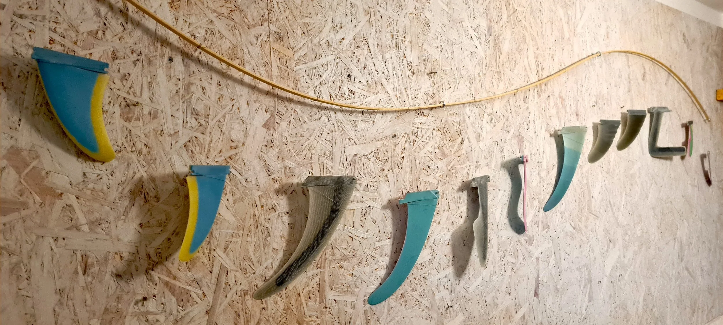 A row of Sam Heaton's fins in varied shapes and colours, some sponge-textured, some smooth resin, strung along a bowed cane rod against a rough plywood wall.