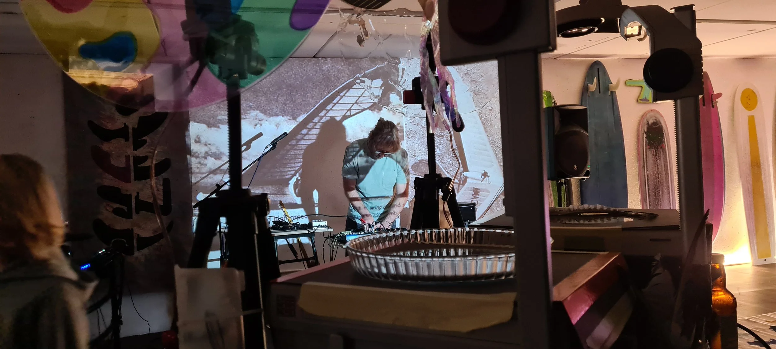 Ken Gunning of Departures at a mixing desk, seen through the coloured discs and projected imagery of a live analog light show, with fin artworks displayed on the wall behind him.