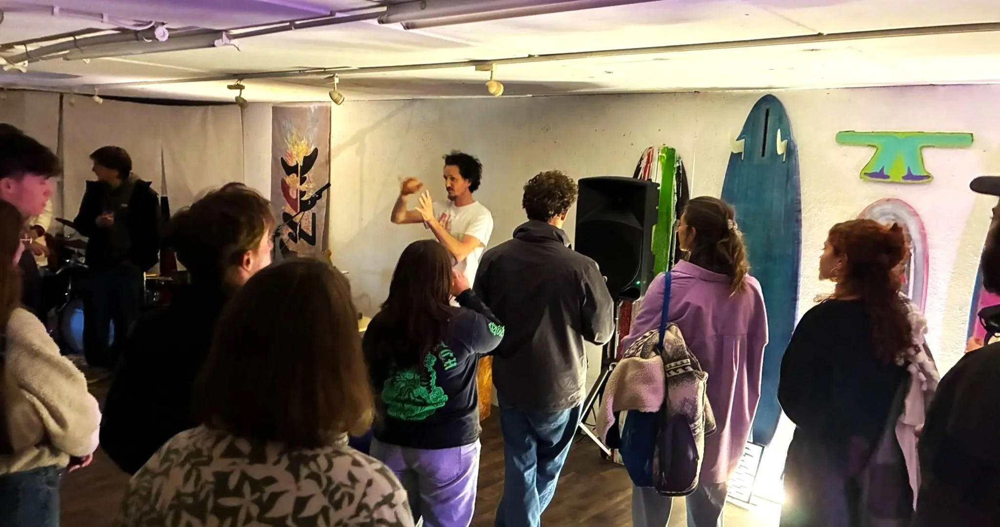 an Jermyn addresses a crowd during the opening of WAKE III, standing in front of fin artworks and a hanging textile piece, with a band setting up behind him.