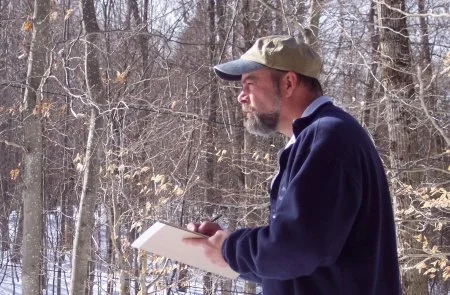 A man with a beard wearing a cap and navy jacket walking through a snowy forest, holding a notebook and pen.