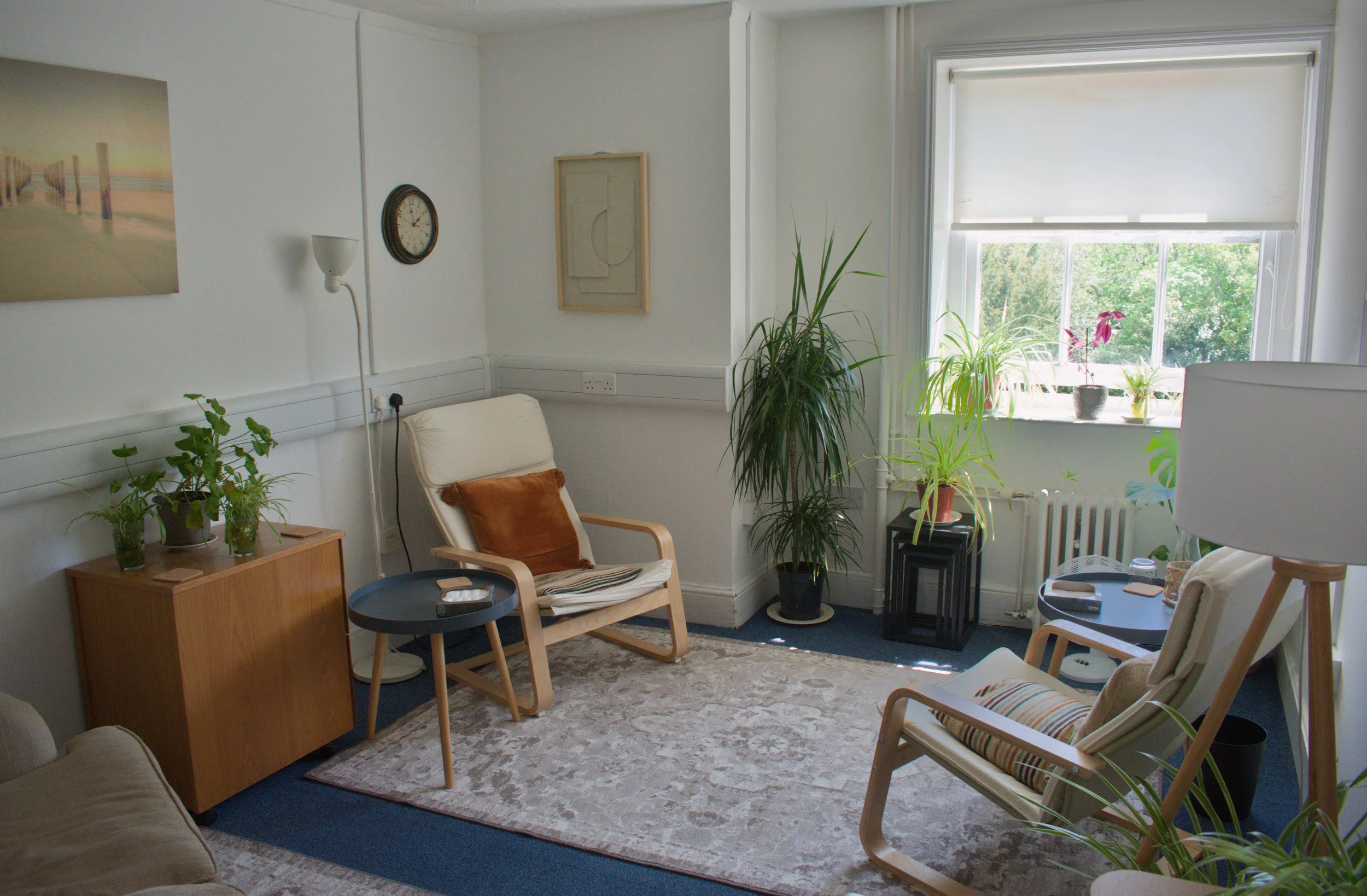 A cozy therapy room with a large window, houseplants, a wooden armchair with pillows, a beige sofa, and artwork on the walls.