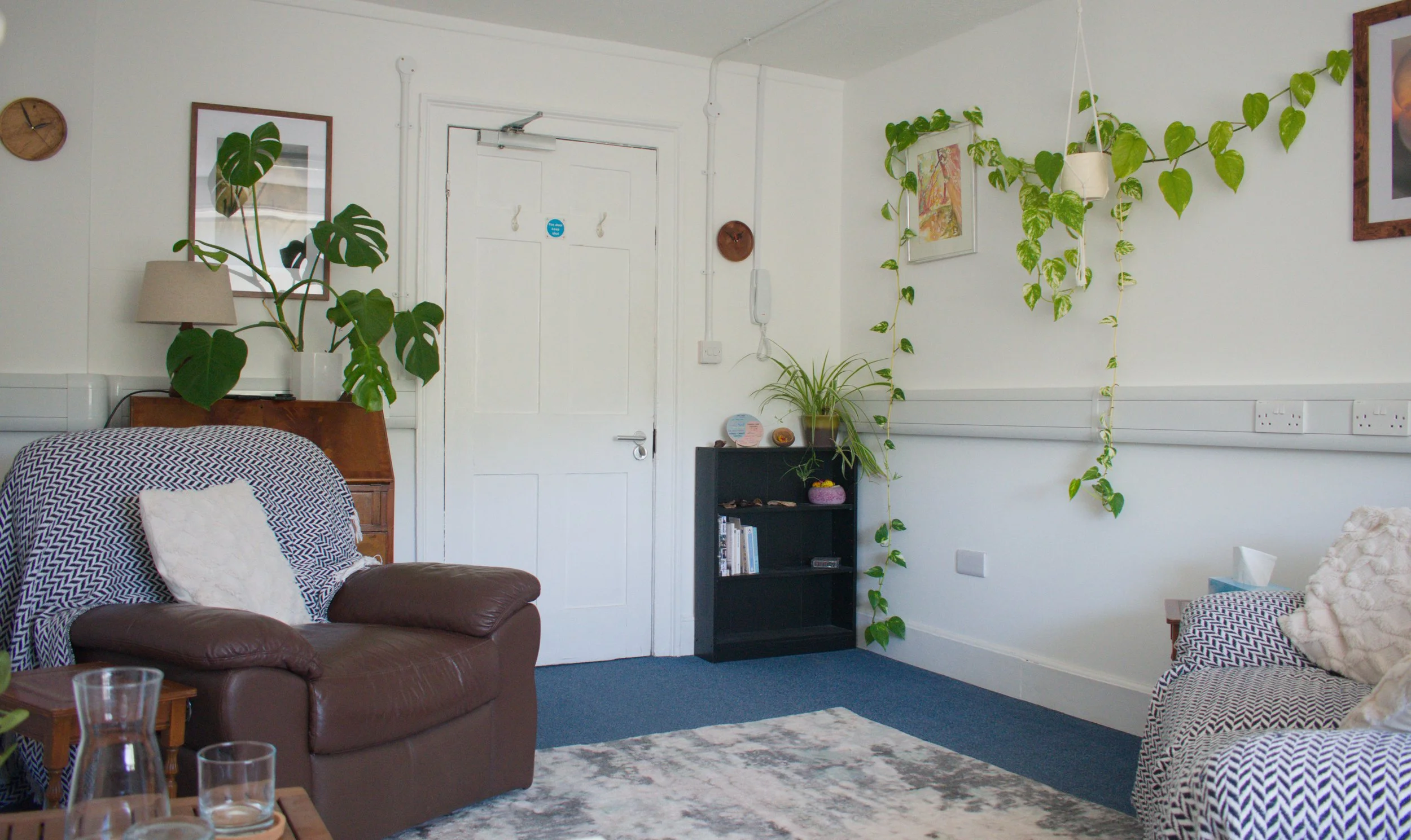 Therapy room with white walls, decorated with pictures, plants, and furniture including a brown leather chair, a dark side table, and a patterned couch. The room has a white door, and a wall clock.