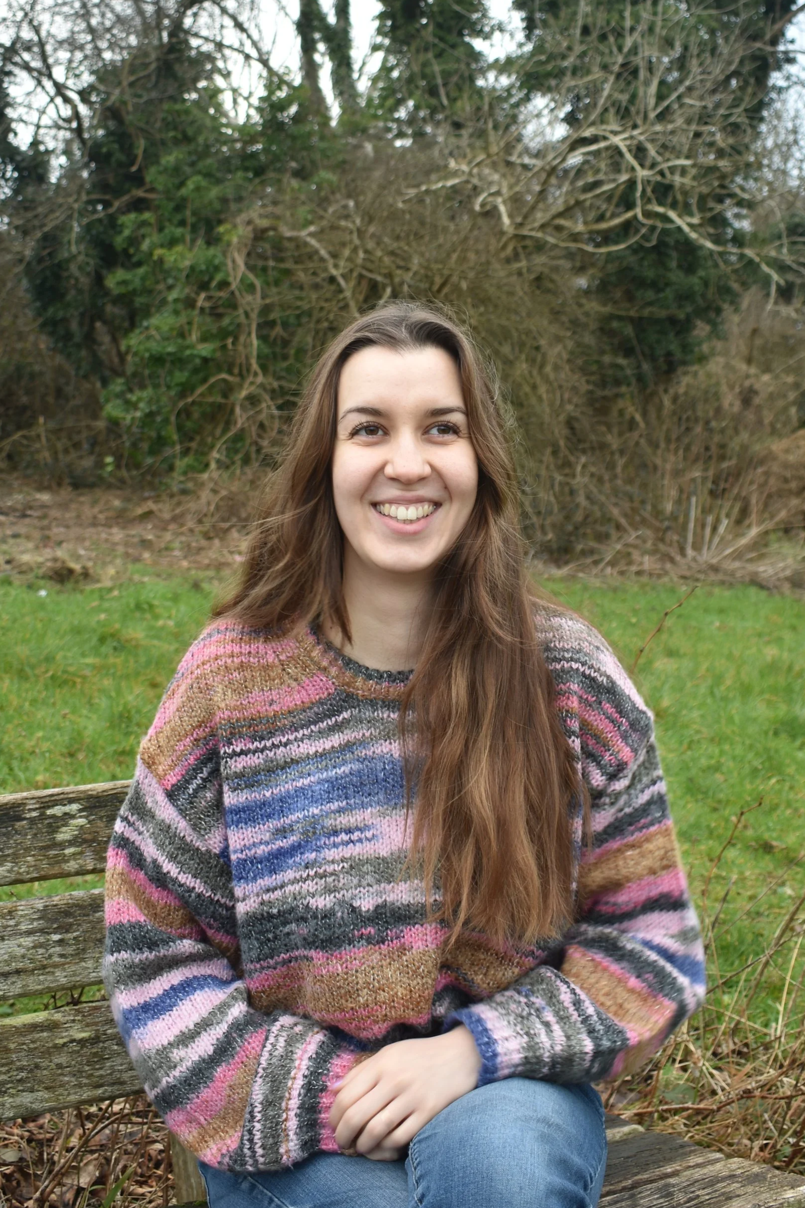 The counsellor (woman with long brown hair) smiling while sitting on a wooden bench outdoors with trees in the background.