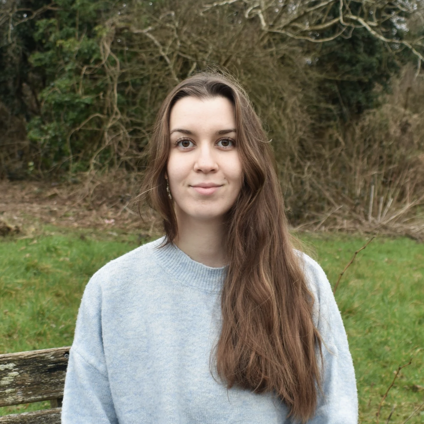The counsellor Milena (woman with long brown hair and light skin) standing outdoors on a grassy area with trees in the background.