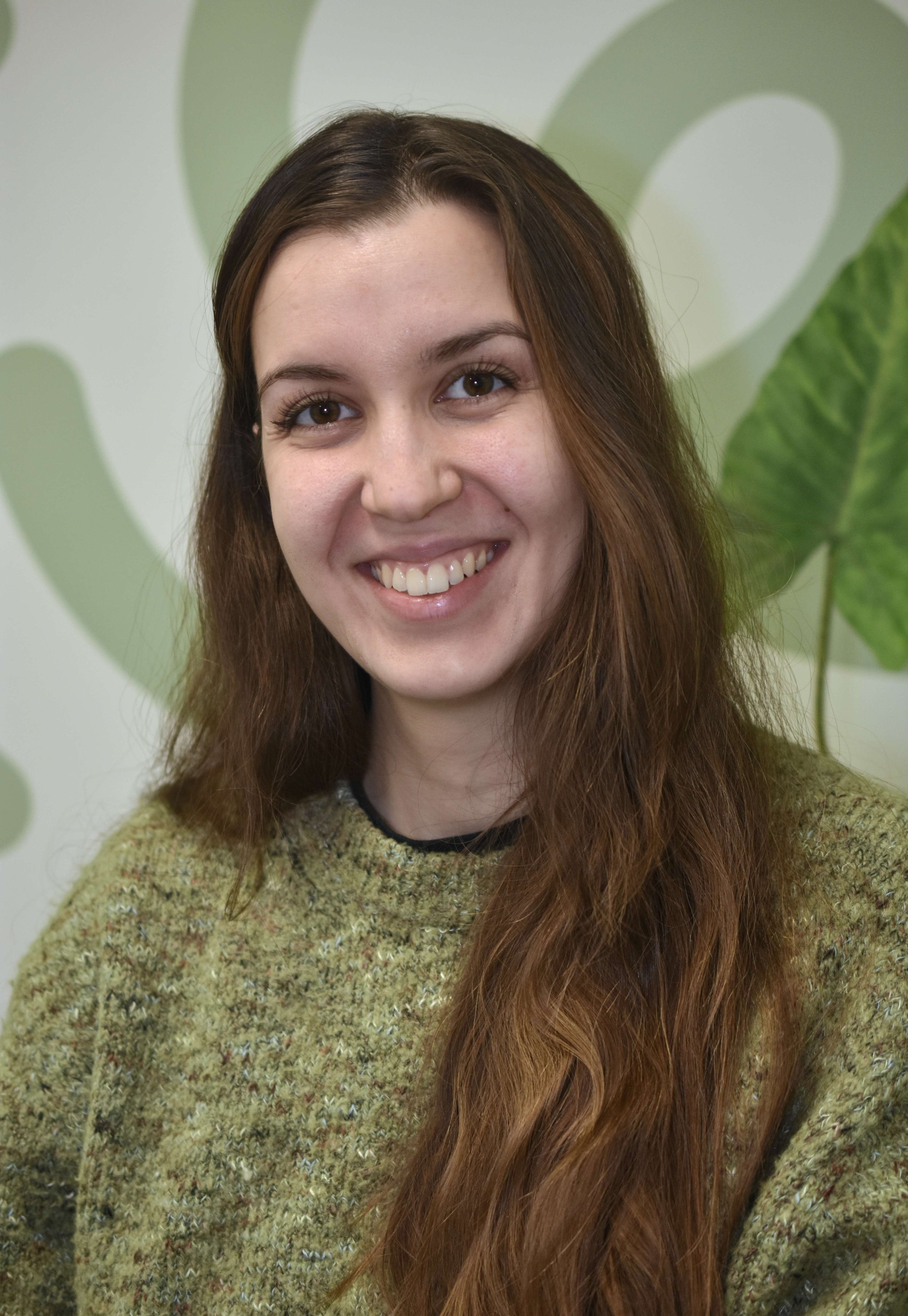 Headshot portrait of therapist Milena Tsankova smiling, with long brown hair and a green textured jumper