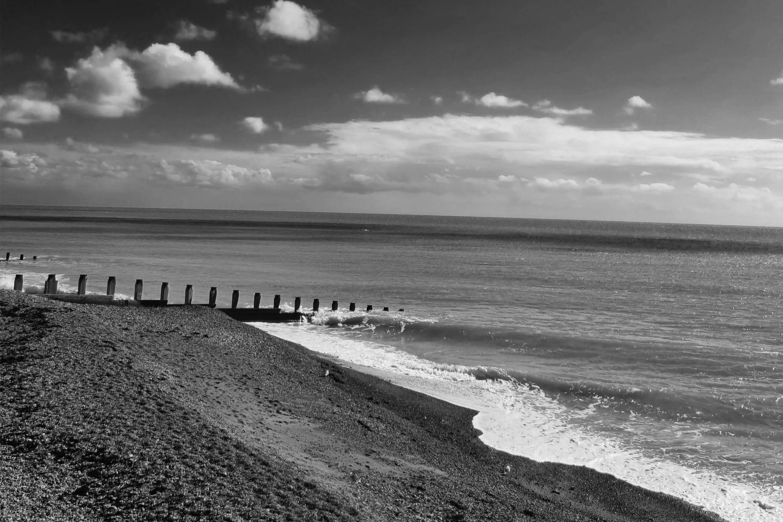 Black and white photo of a beach with a cloudy sky, a shoreline with gentle waves, and a series of wooden posts along the beach.