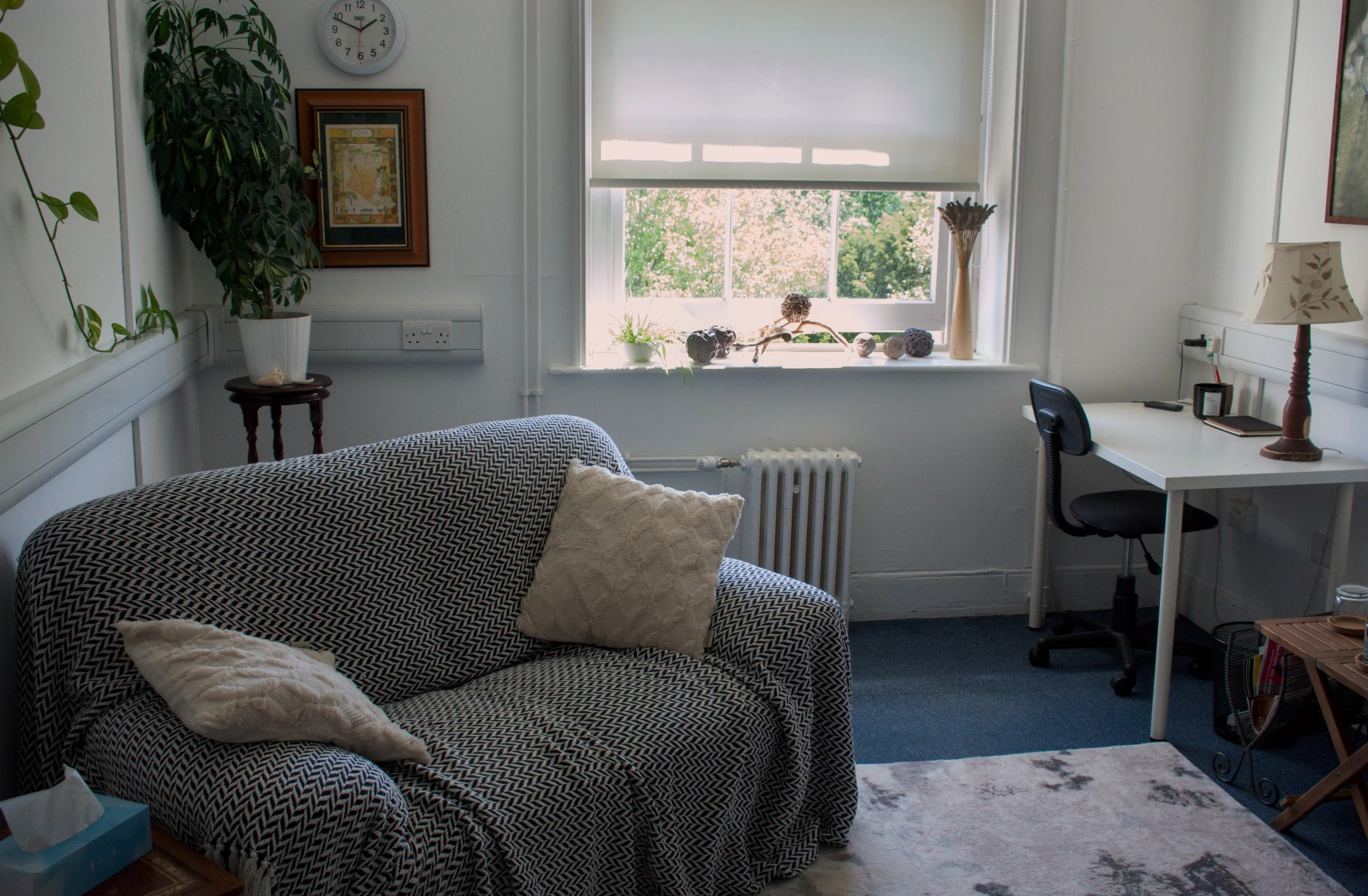 A cozy therapy room with a patterned sofa, a large window with a view of trees, a potted plant, a clock, framed artwork, and a small desk with a lamp and chair.