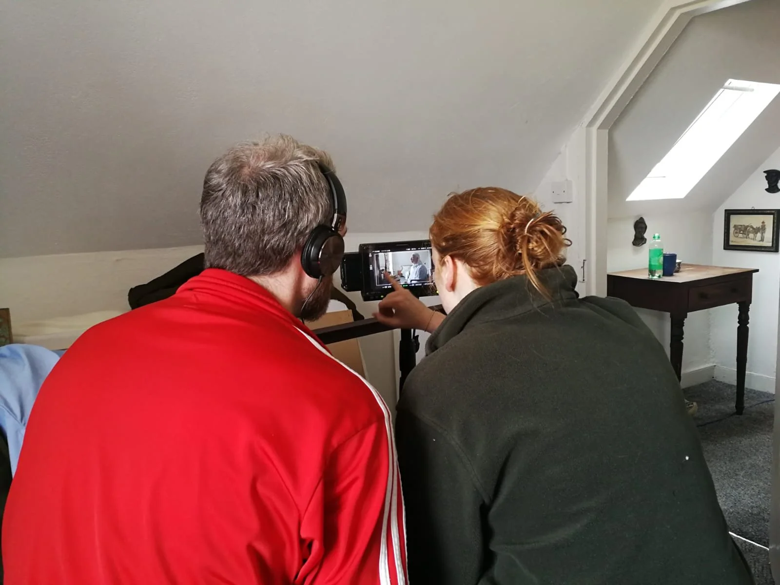 A behind the scenes photo. Two people sitting on a bed, one wearing headphones and the other pointing at a video monitor.