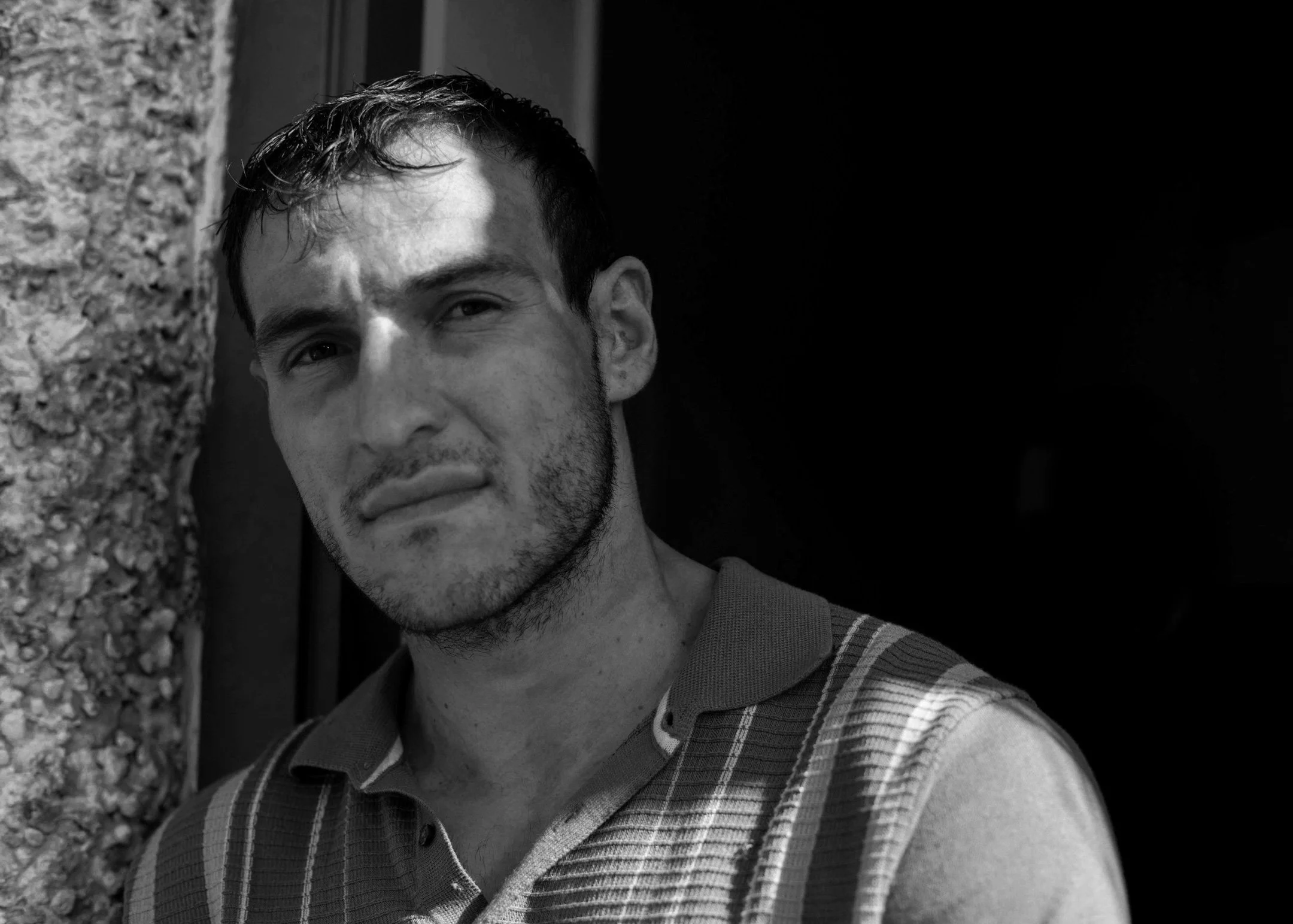 A young man with short dark hair, light stubble, wearing a plaid shirt, stands against a stone wall and dark background.