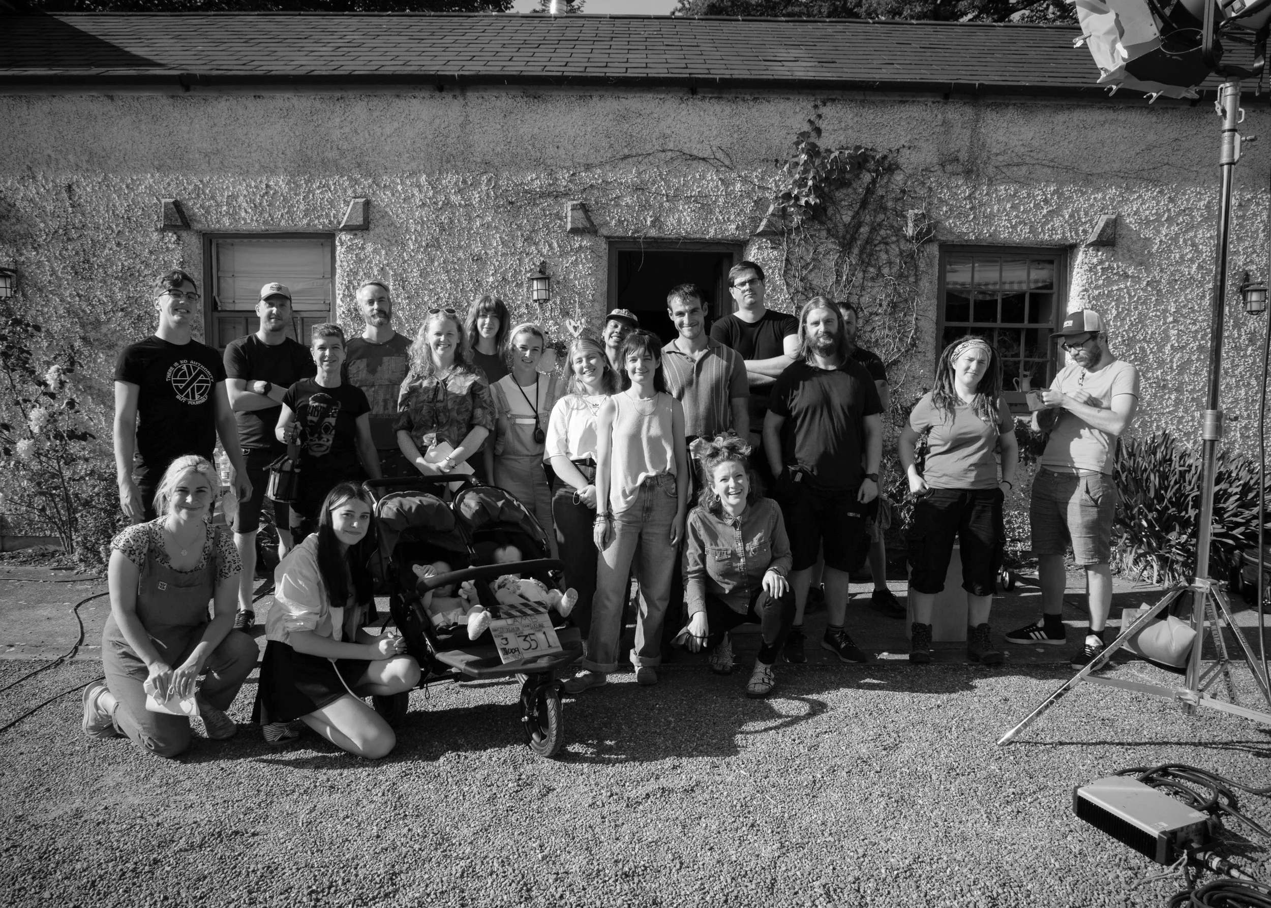 A group of people posing outside in front of a textured house wall with windows, climbing plants, and outdoor lights. The group includes adults and children, some smiling and gathered around a stroller. Camera and lighting equipment are visible on th