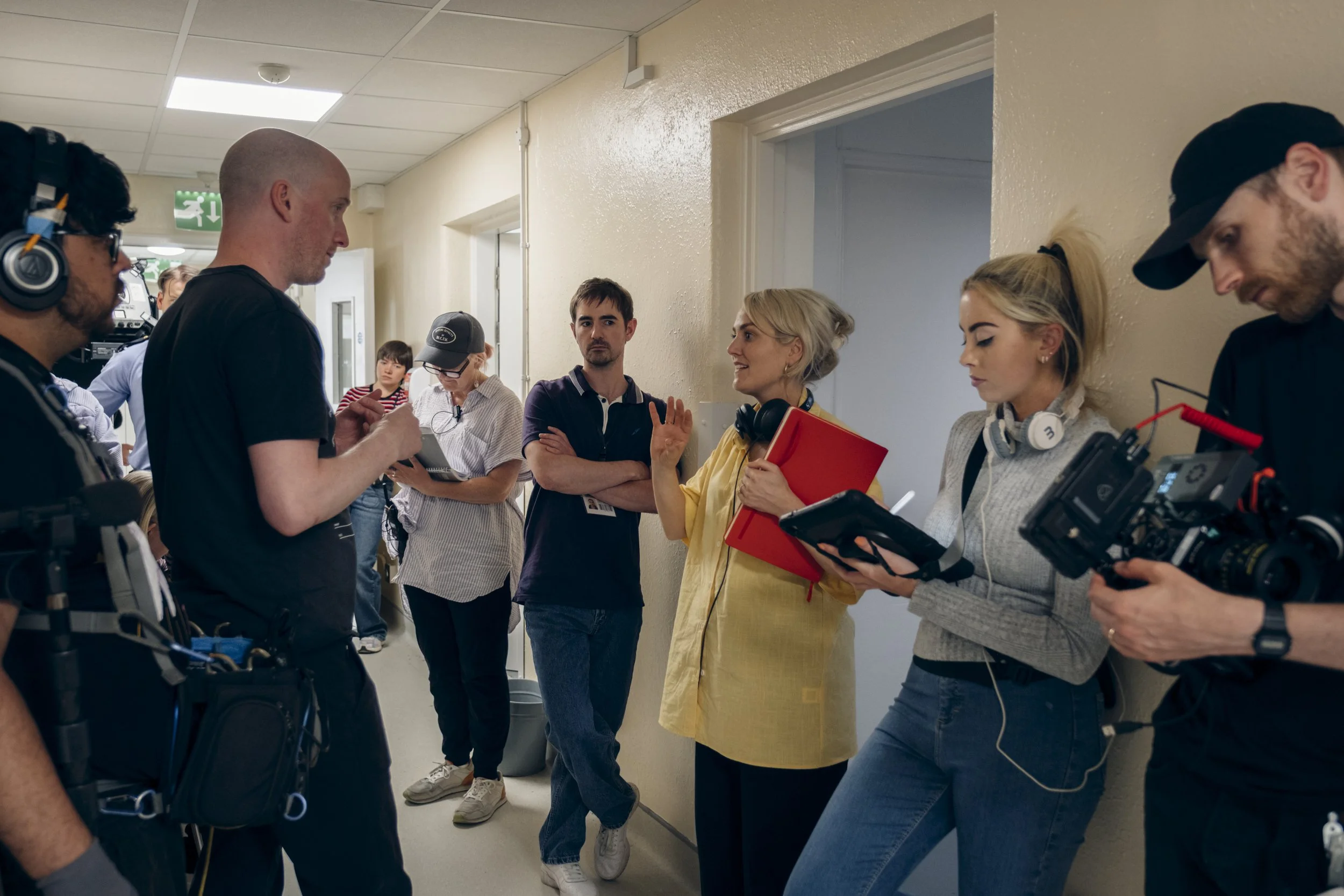 Group of people standing and talking in a hallway with one woman gesturing while holding a red folder. Several individuals are wearing headphones or microphones, and some have equipment, suggesting a film or video production setting.