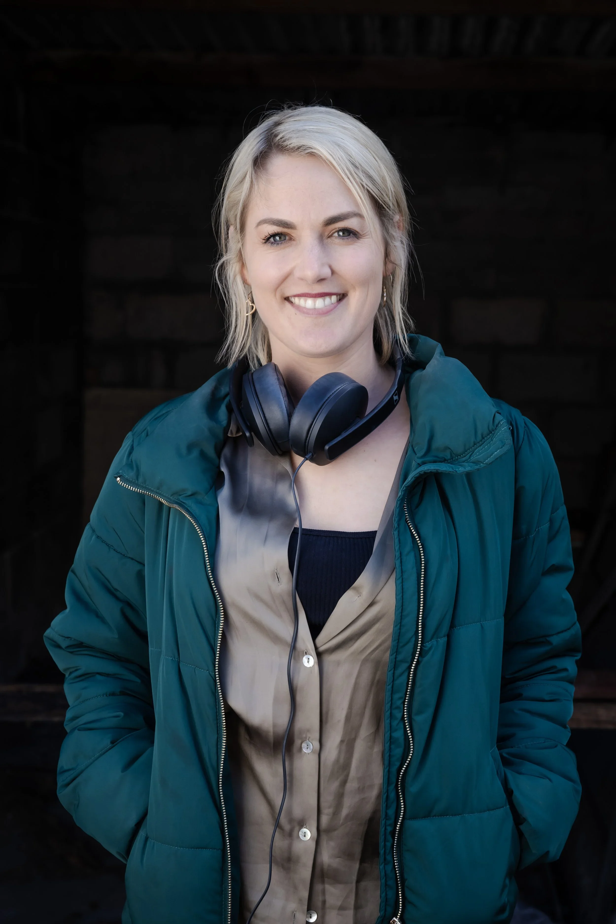Smiling woman with short blonde hair wearing a teal jacket and headphones around her neck, standing outdoors.