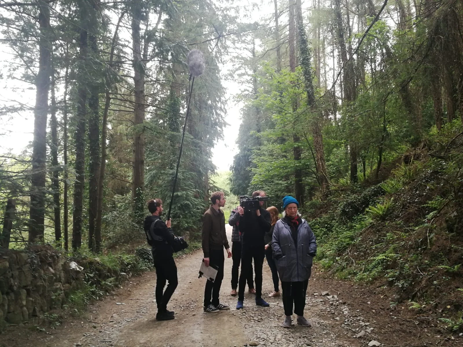 A group of people, including a camera operator, are standing on a forest trail. One person is holding a boom microphone, and some are talking or observing the surroundings.