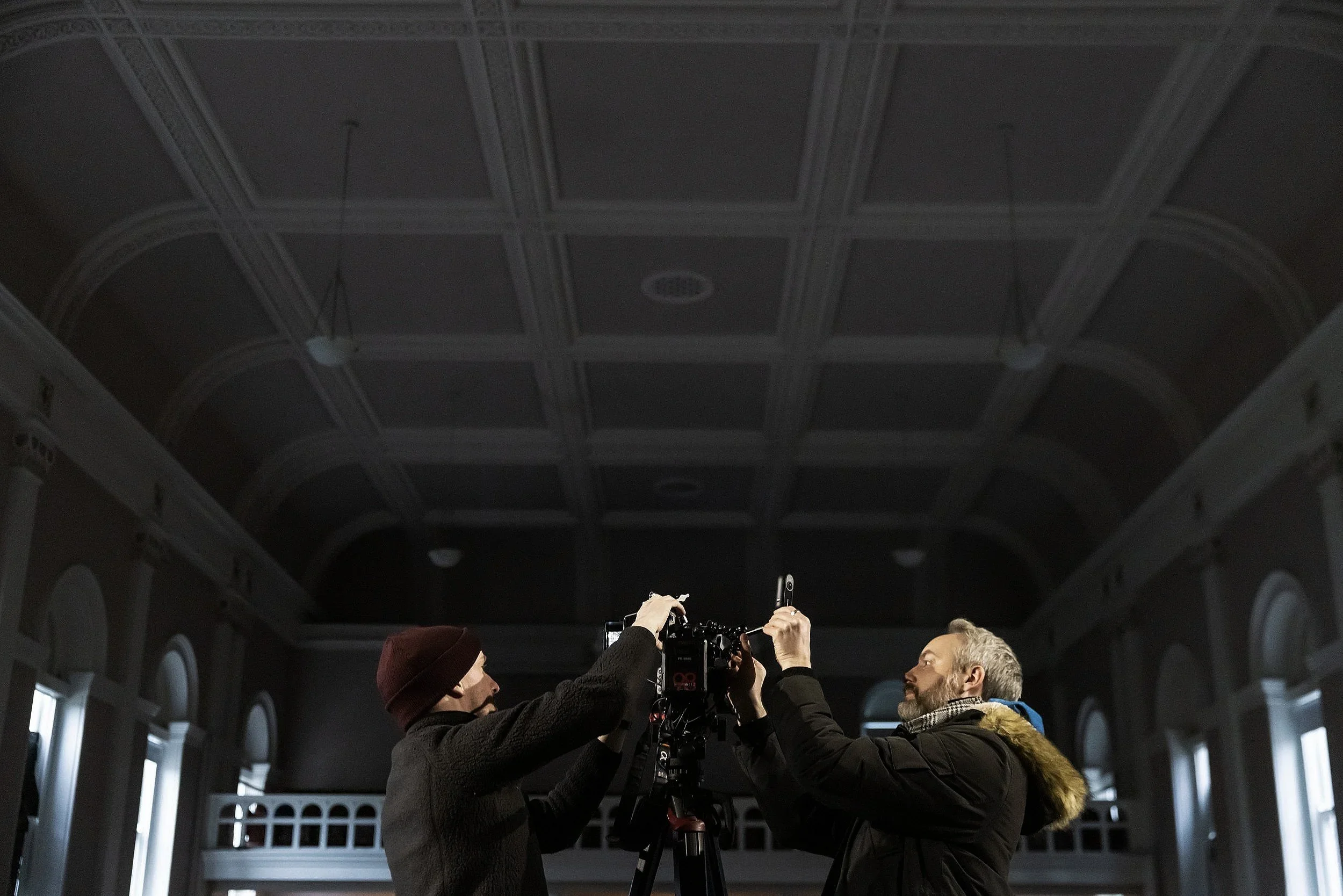 Two men setting up a camera in a large, dimly lit room with high vaulted ceiling and tall windows.