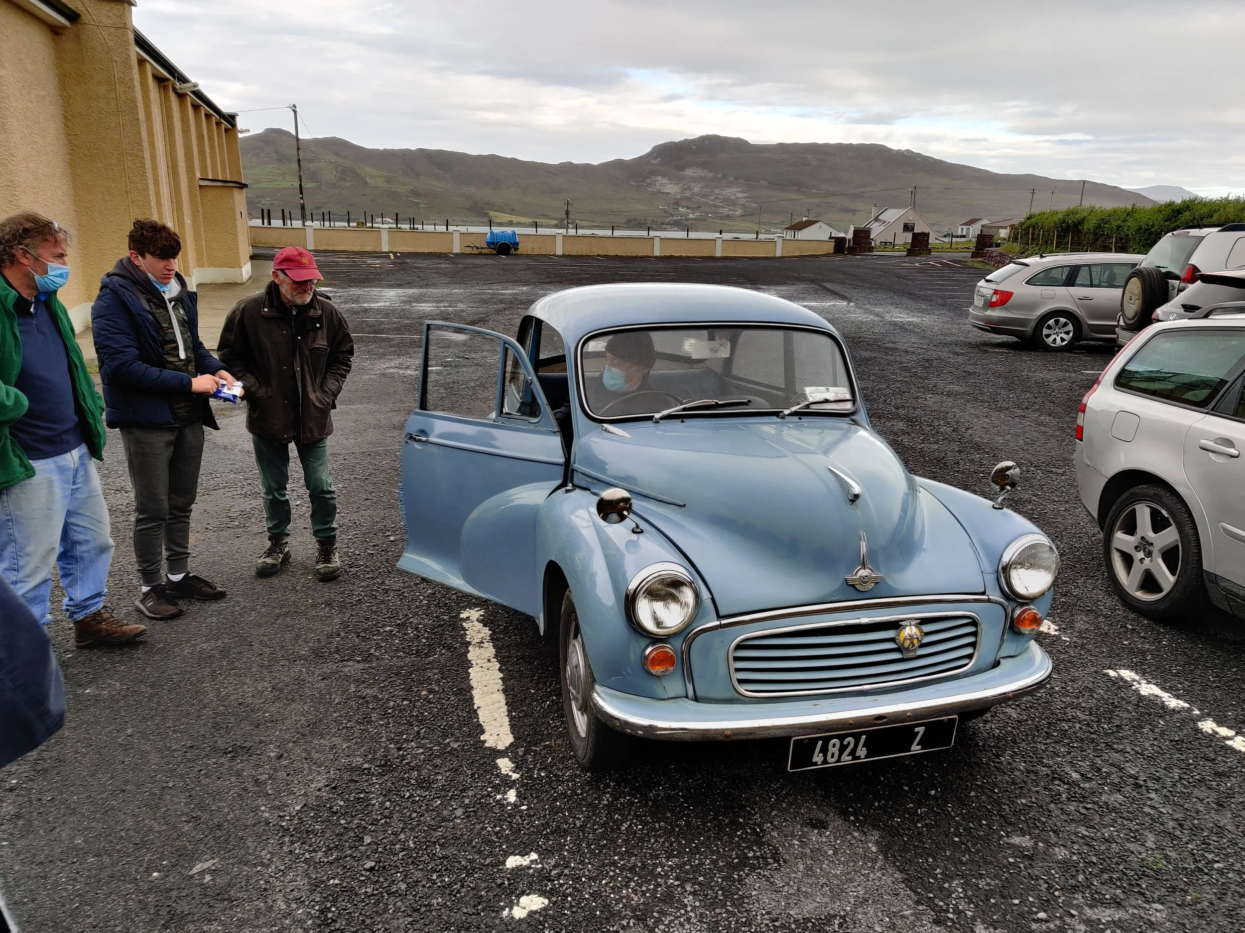 Behind the scenes photo.A scenic parking lot with a vintage blue microcar in the foreground, surrounded by modern cars, with mountains and cloudy sky in the background. Three men wearing face masks are near the microcar, one holding a remote control.