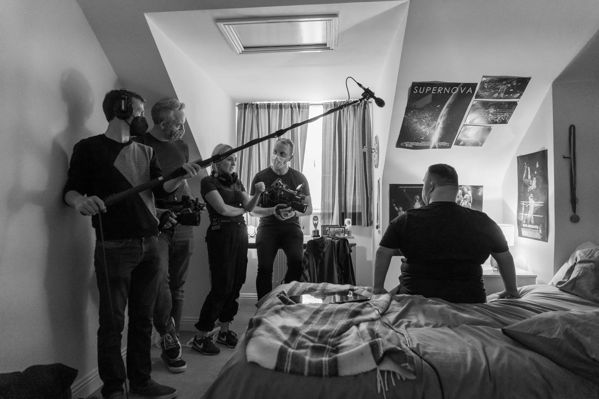 Group of five people in a bedroom, filming or conducting an interview with a third person seated on the bed. The person on the bed has short hair and is wearing a black shirt. The four standing individuals are wearing face masks, one is holding a boo