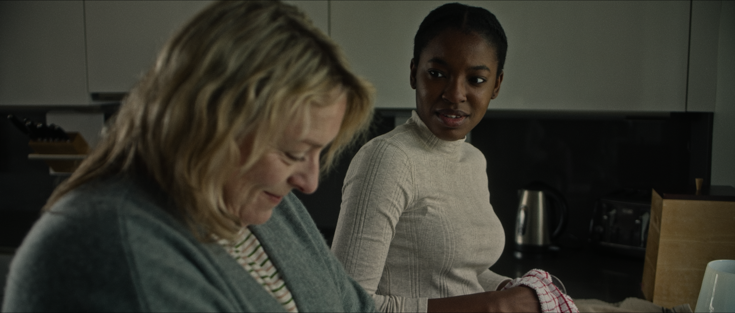 Two women in a kitchen; one with blonde hair looking down, smiling, and wearing a striped shirt and green sweater, the other with dark hair in a bun, looking at her, wearing a beige turtleneck